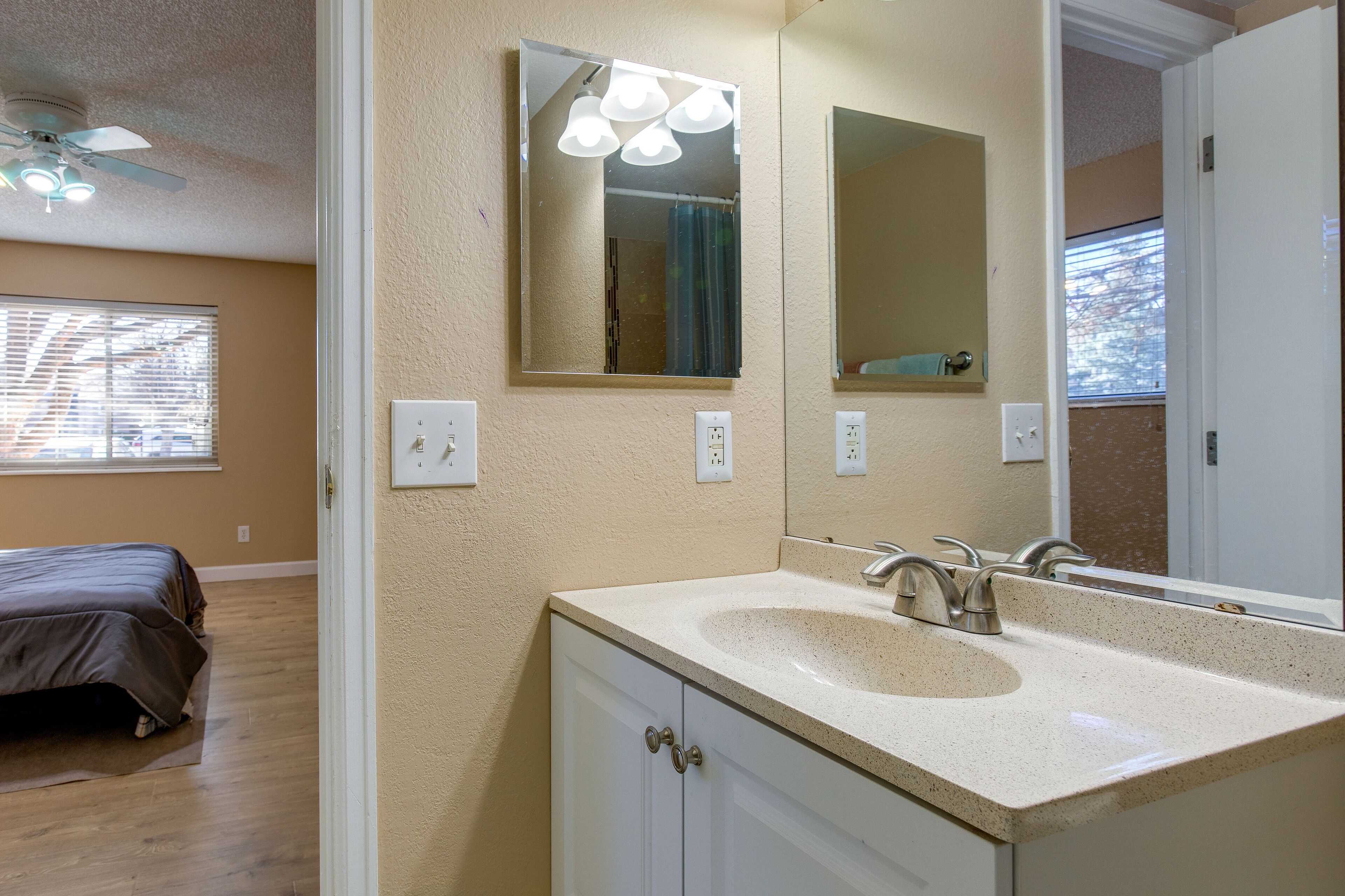 Une salle de bain avec un comptoir beige, un lavabo et un miroir. Un ventilateur de plafond est installé au-dessus du lavabo, et une fenêtre avec des stores se trouve à l’arrière-plan.