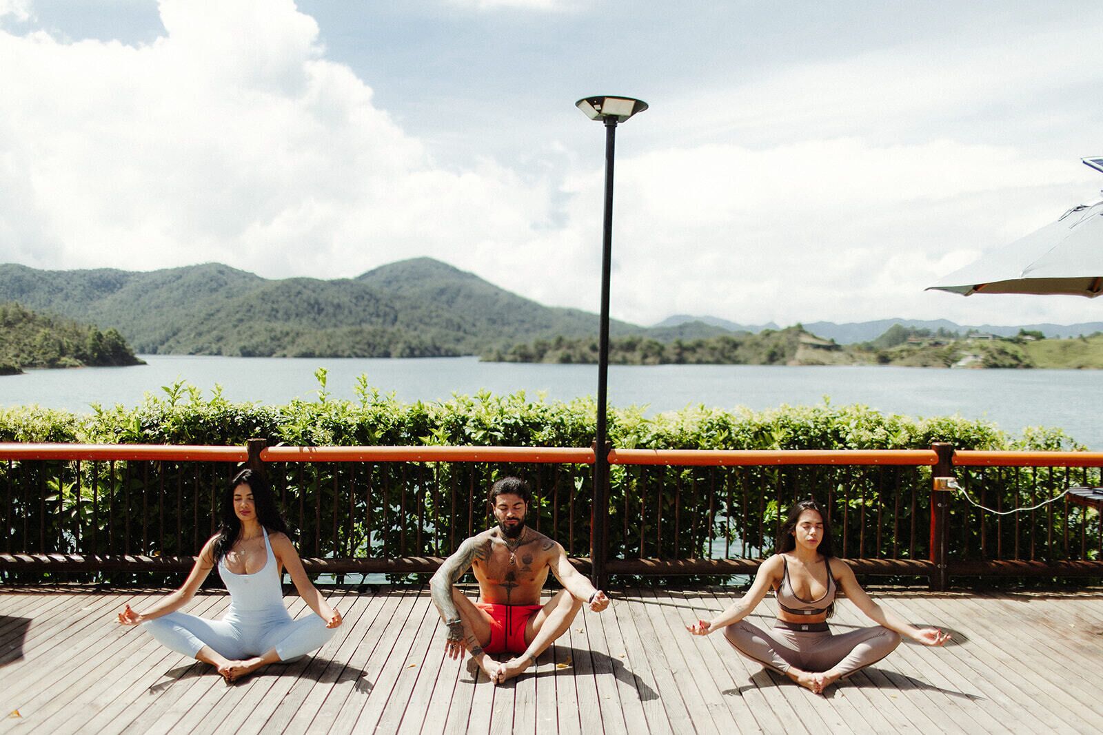 Três pessoas meditando em um deck de madeira, com vista para um lago e montanhas.