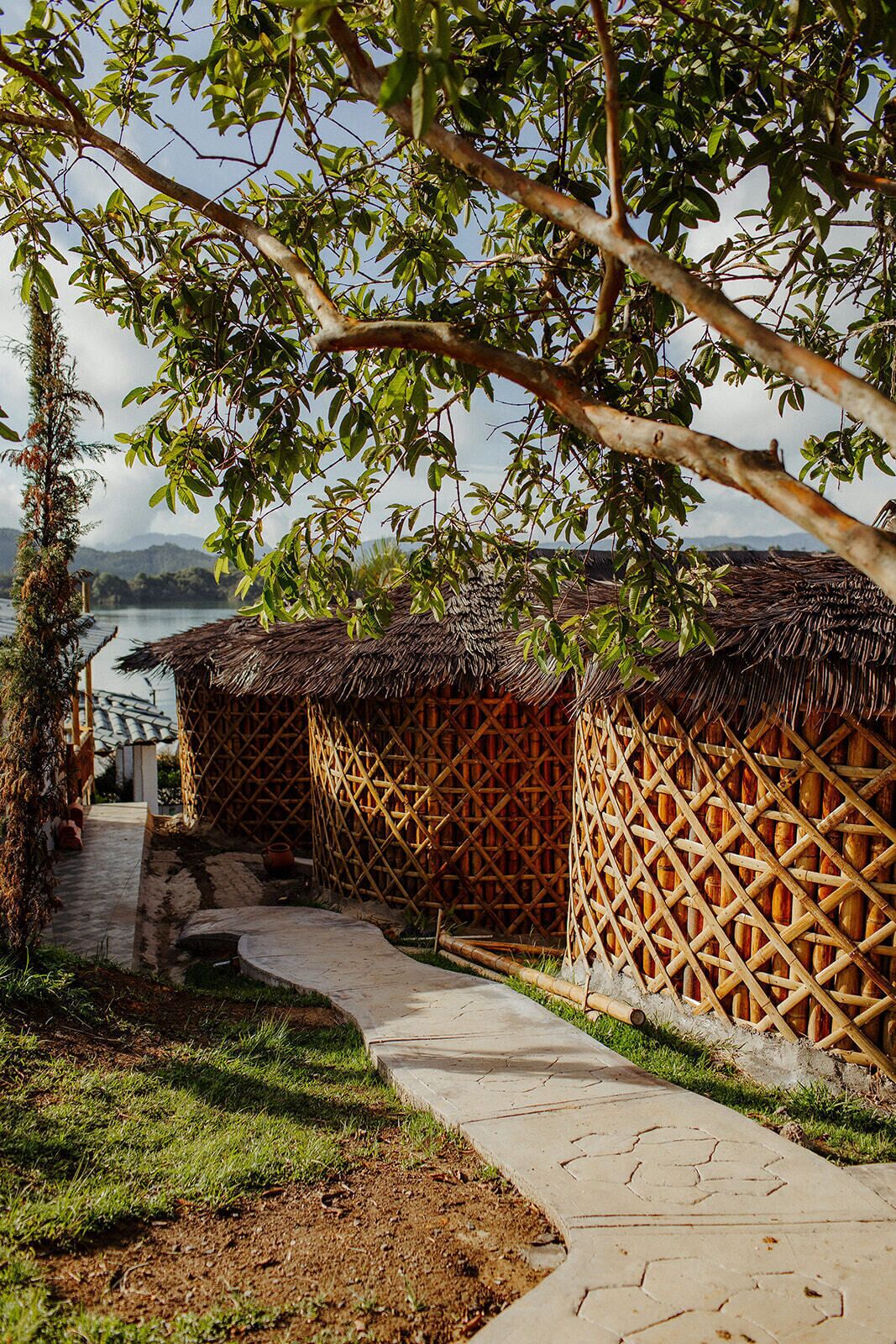 A pathway leading to thatched-roof huts with a view of a body of water and mountains in the background.
