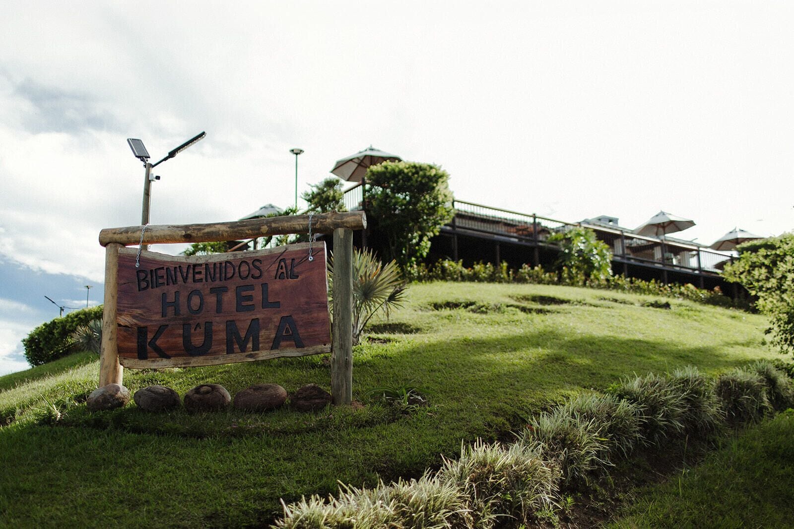 A wooden sign with 'BIENVENIDOS AL HOTEL KUMA' text, a solar panel, and a building with a balcony in the background.