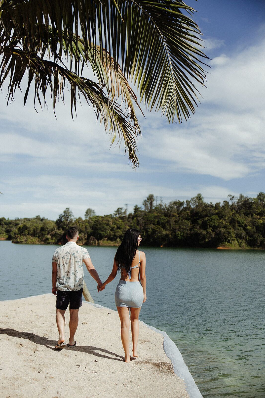 A couple holding hands on a sandy beach with a body of water and trees in the background.