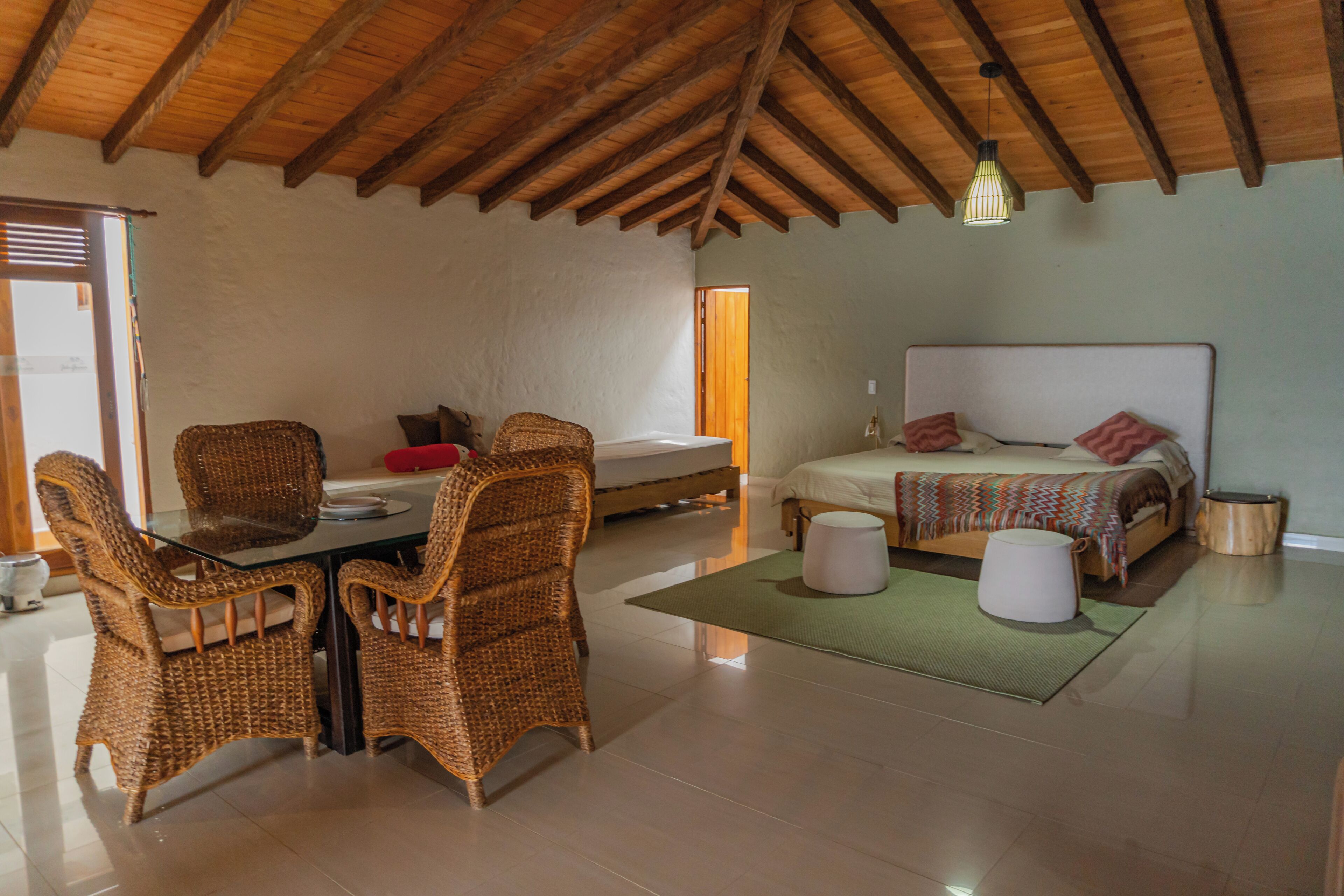 A room with a high ceiling, a bed with a patterned bedspread, a dining table with wicker chairs, and a small round table with a lamp.