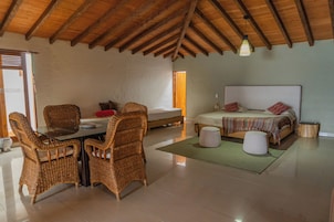 Una habitación con techo alto, una cama con colcha estampada, una mesa de comedor con sillas de mimbre y una mesita redonda con lámpara.