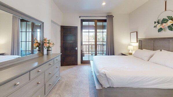 A hotel room with a large bed, a dresser with drawers, a mirror, a door leading to a balcony, and a wall-mounted lamp.