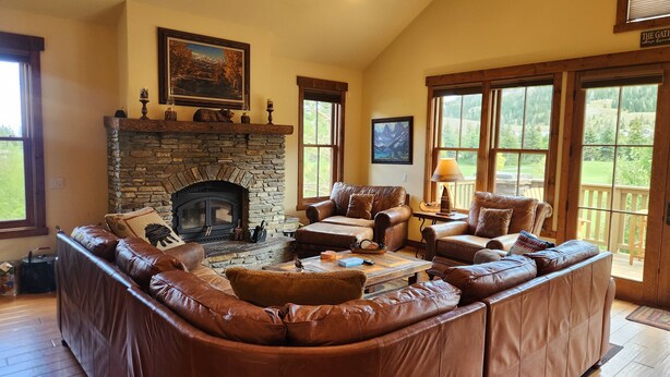 A cozy living room with a stone fireplace, leather sofas, and large windows offering a view of the outdoors.