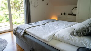 A bedroom with a bed, a bedside table, a nightstand, and a window with a view of greenery.