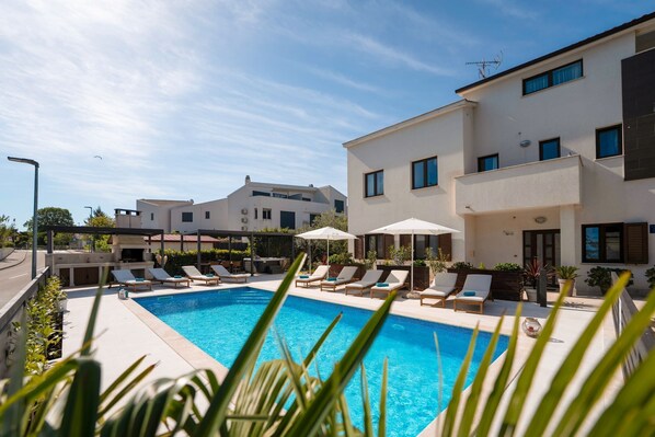 Una piscina con sillones de descanso y sombrillas, rodeada por un edificio moderno con múltiples ventanas y balcones.