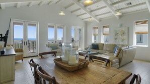 A spacious living area with a wooden dining table, wicker chairs, a leather sofa, and a large window offering a view of the ocean.