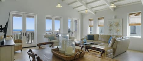 A spacious living area with a wooden dining table, wicker chairs, a leather sofa, and a large window offering a view of the ocean.