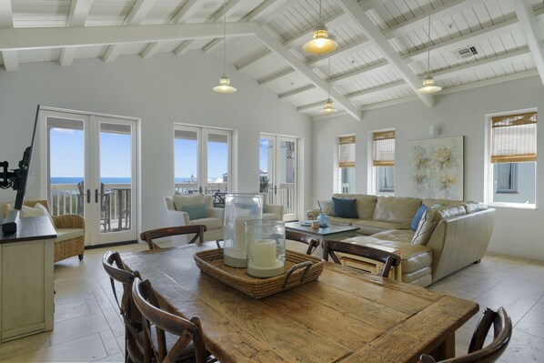 A spacious living area with a wooden dining table, wicker chairs, a leather sofa, and a large window offering a view of the ocean.