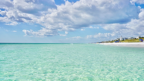A beach with clear turquoise water, a sandy shore, and buildings in the background.