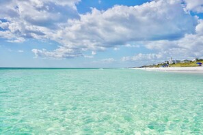 A beach with clear turquoise water, a sandy shore, and buildings in the background.