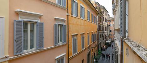 A narrow street with buildings on either side, featuring multiple windows with shutters, a cobblestone path, and pedestrians.