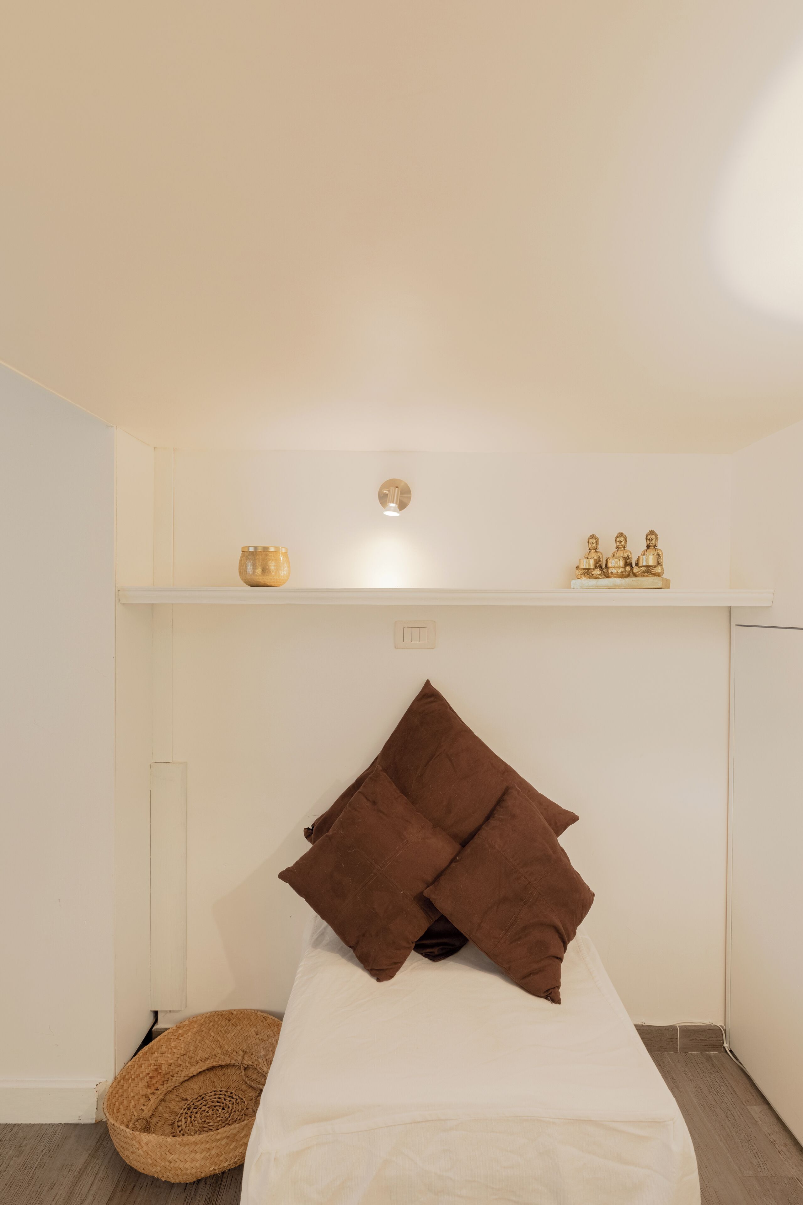 A bedroom with a bed, a brown pillow, a small wooden bowl, and a shelf with three golden Buddha statues.