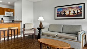 A modern kitchen with wooden cabinets, a microwave, and a bar stool. There is a gray sofa and a small wooden table in the living area. - 1 Bedroom Condo :: Oceanfront Resort Kapaa Shore (Kauai)