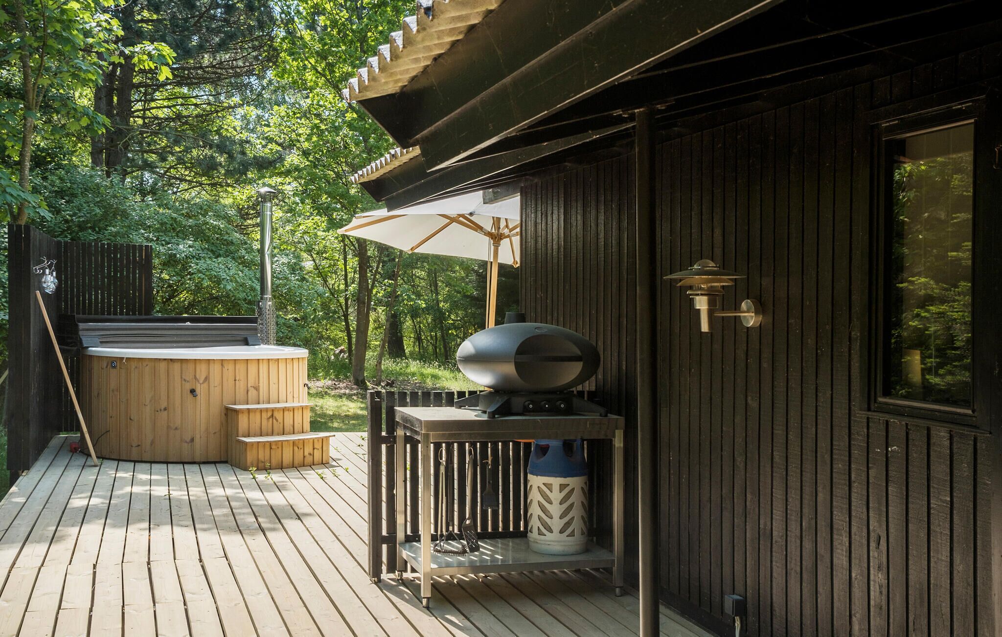 Une terrasse en bois avec un jacuzzi, un barbecue et une pergola dotée d’un parasol.