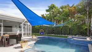 A swimming pool with a blue shade sail, a glass-enclosed patio area with outdoor furniture, and a lush green hedge in the background.