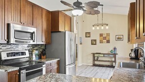 A kitchen with wooden cabinets, a stainless steel refrigerator, a microwave, a stove, granite countertops, a ceiling fan, and a small dining area with a table and chairs.