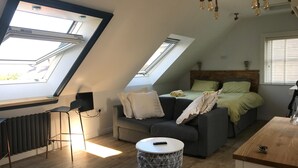 Ein kompakter, moderner Raum mit schrägverdeckter Decke, Dachfenstern, einem Bett mit grüner Bettwäsche, einem grauen Sofa und einem kleinen runden Tisch.