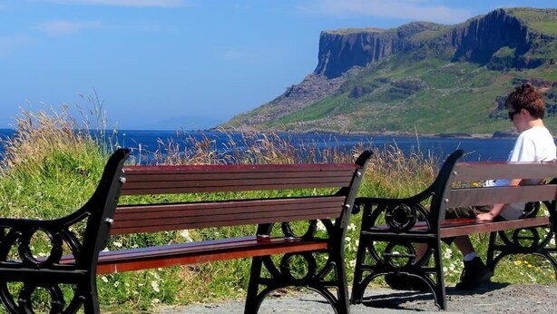 Eine Bank mit einer sitzenden Person, mit Blick auf eine Küstenlandschaft mit Klippen und dem Meer.