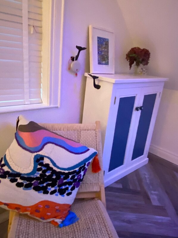 A room with a white chair, a colorful blanket, a white cabinet with a picture and a vase, and a window with blinds.