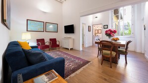 Een woonkamer met een blauwe bank, rode stoelen, een houten eettafel en een televisie aan de muur.