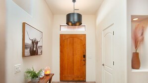 A hallway with a wooden door, a hanging light fixture, a framed picture, a small table with a plant and candle, and a white door on the right. - Enchanted Adobe Hideaway w/Hot Tub & GIANT Chess (Sedona)