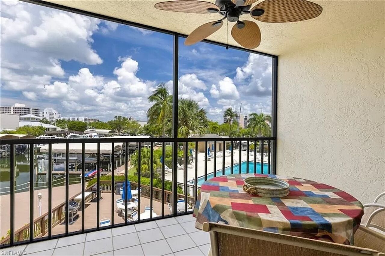 A balcony with a view of a swimming pool and palm trees, a ceiling fan, and a table with a colorful tablecloth.