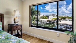 A bedroom with a large window overlooking a marina, a bed with a floral bedspread, and a nightstand with a lamp.
