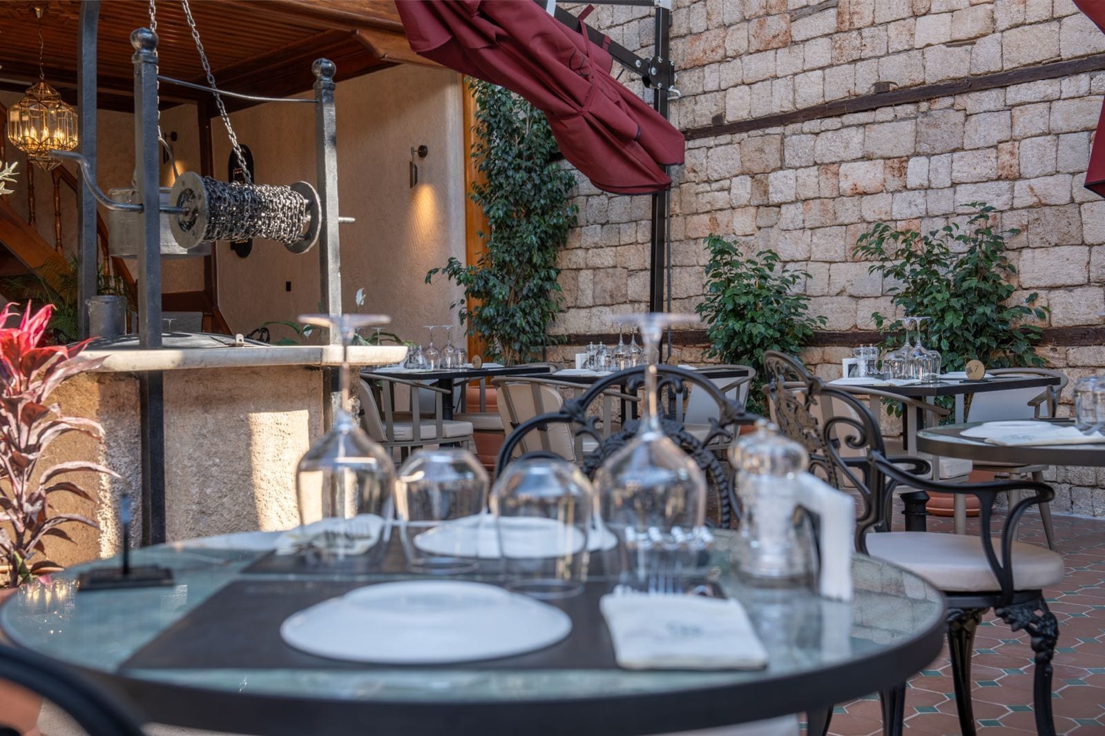 An outdoor dining area with tables set for two, featuring metal chairs and a stone wall backdrop.