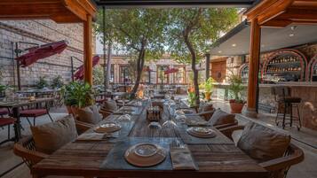 An outdoor dining area with wooden tables and chairs, surrounded by stone walls and potted plants.