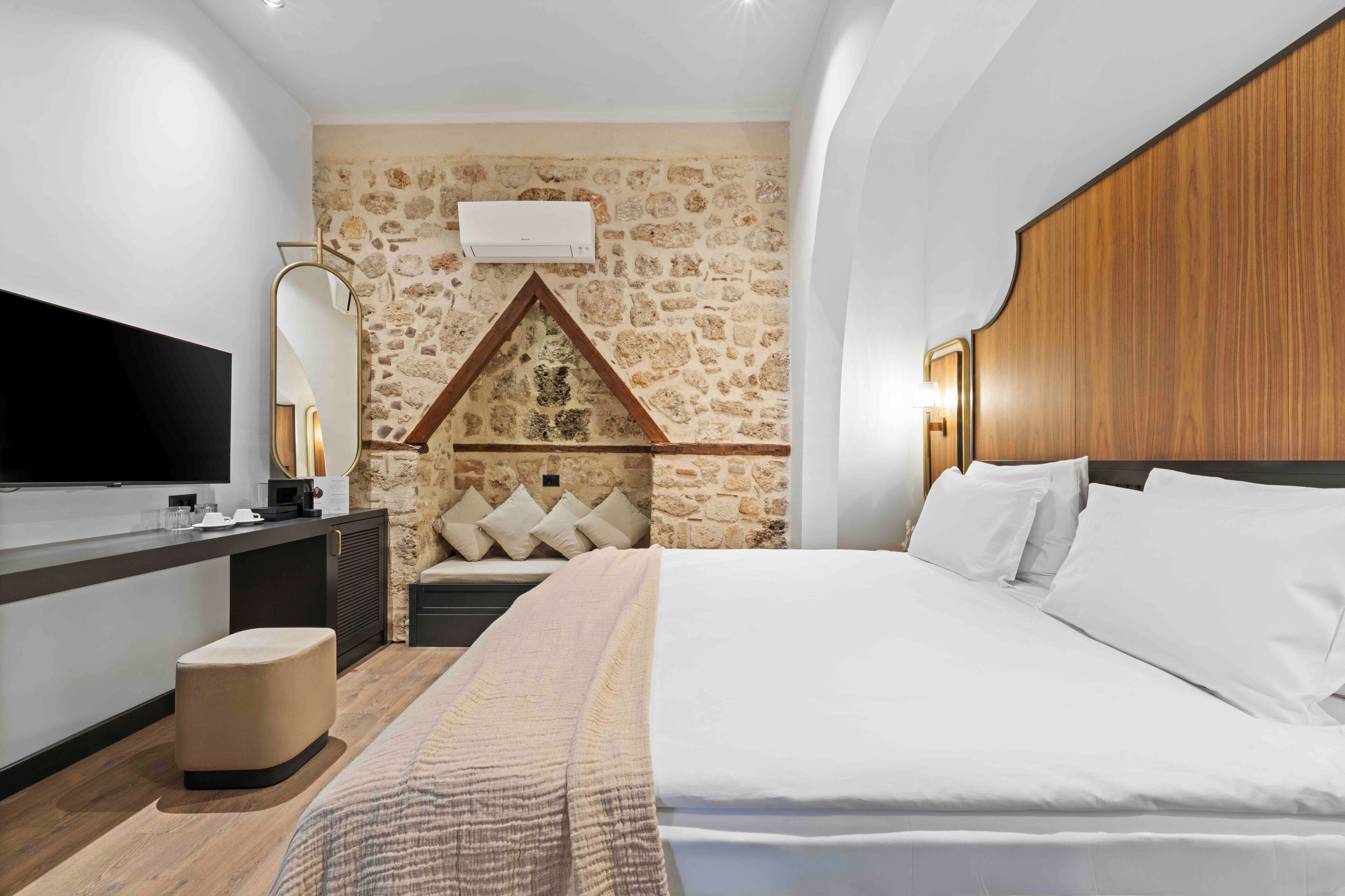 Un dormitorio con pared de piedra, cabecero de madera, televisor de pantalla plana, mesita de noche y cama con ropa blanca.
