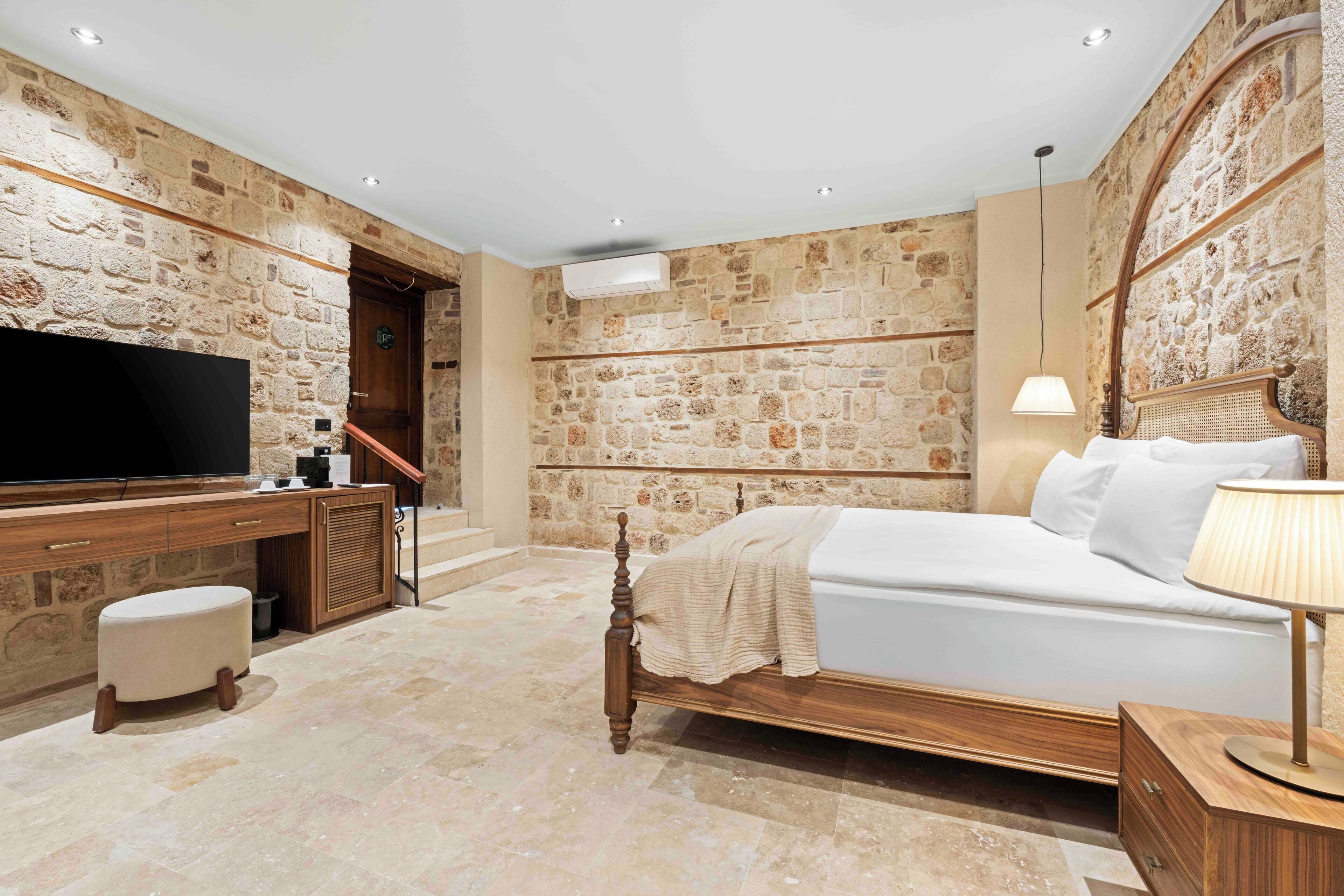 A bedroom with a stone wall, a bed, a TV, a desk, and a lamp.