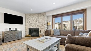 A living room with a stone fireplace, a sofa, a coffee table, and a TV mounted on the wall. - Perfectly Located 2 Minute Walk Lift Downtown Avon (Avon)