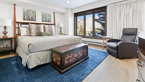A bedroom with a large bed, a wooden chest, a grey armchair, and a window with a view of snow-covered trees. - Perfectly Located 2 Minute Walk Lift Downtown Avon (Avon)
