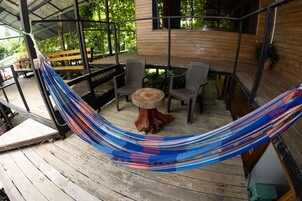 Una hamaca en una terraza de madera con una mesa y sillas, rodeada por una estructura de madera con ventanas.