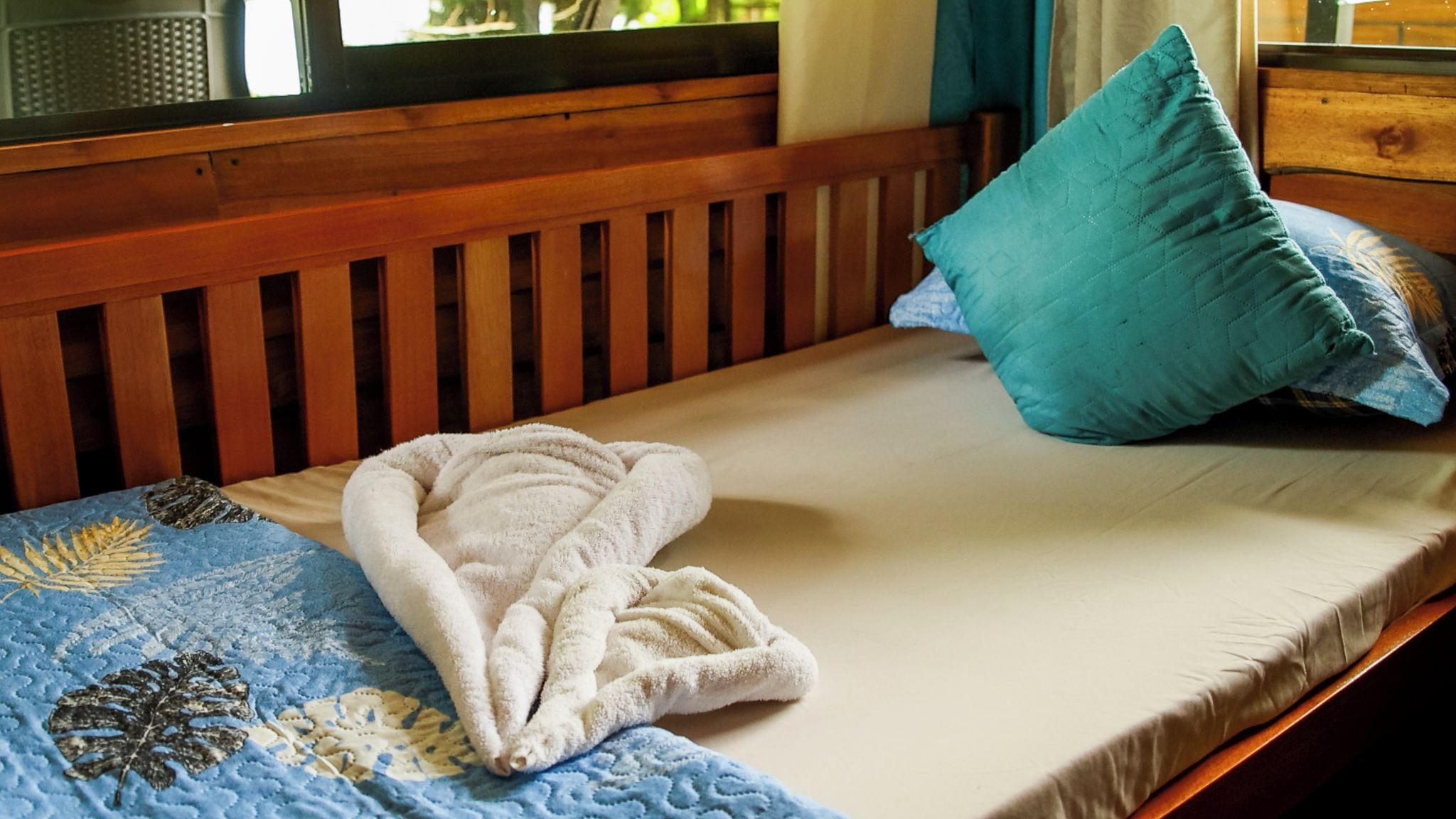 A bed with a blue quilt, a white towel, and a teal pillow.