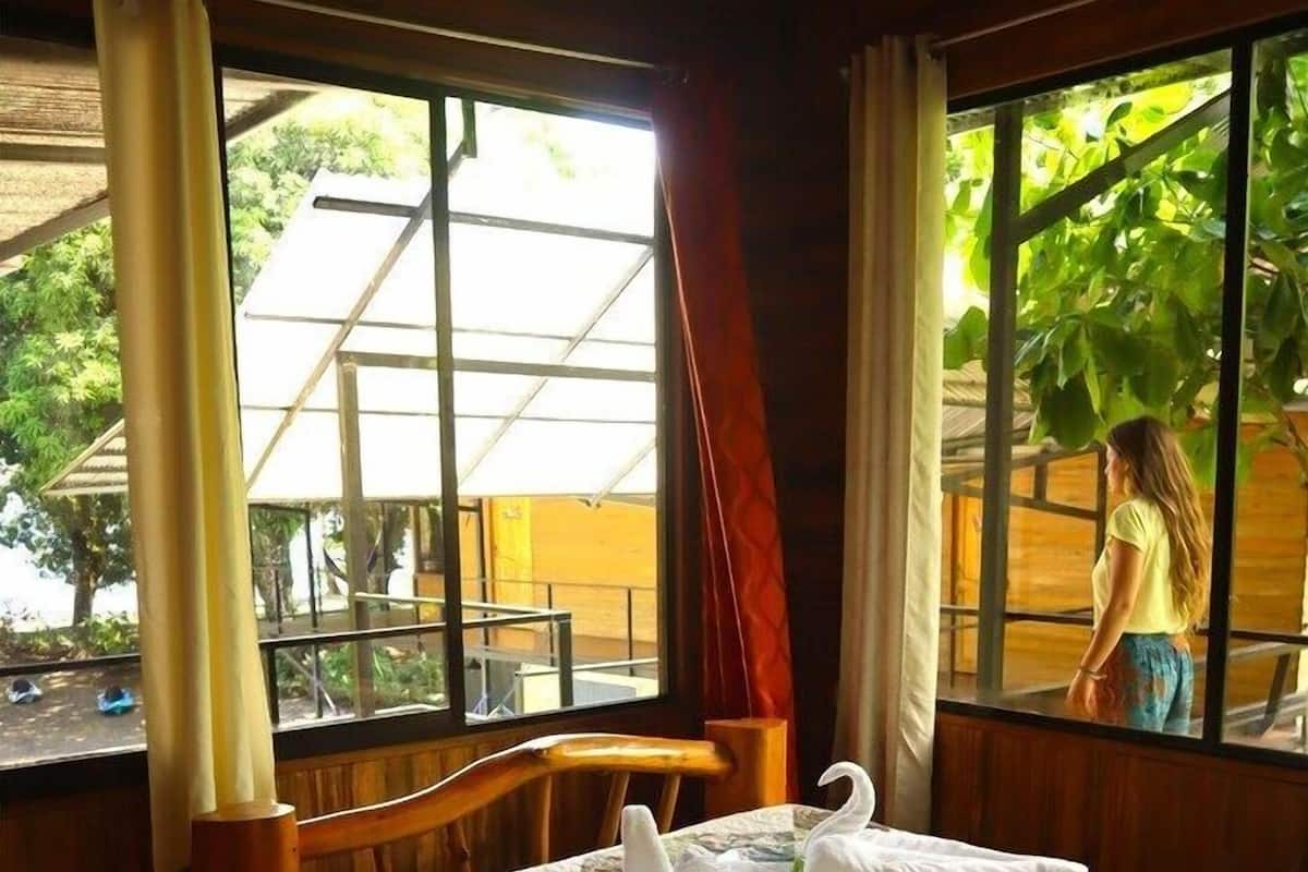 A wooden-framed room with a bed, two windows, and a view of a person outside.