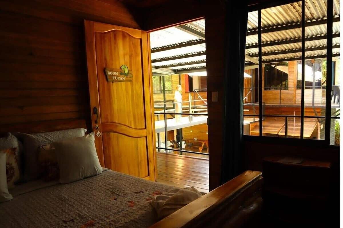 A wooden room with a bed, a sign reading "ROOM TUCAN," and a view of an outdoor area with tables and chairs.