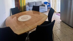 A dining area with a wooden table and black chairs, a grey sofa, and a surfboard leaning against the wall. - Apartamento Centro de Jaco, 100 Metros Playa, Comercios (Jacó)