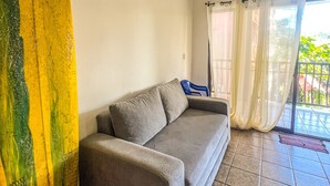 A living room with a grey sofa, a surfboard leaning against the wall, and a view of the outdoors through a glass door. - Apartamento Centro de Jaco, 100 Metros Playa, Comercios (Jacó)