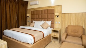 A hotel room with a large bed, a wooden headboard, two bedside tables, a chair, and an air conditioning unit. - Wingate Exotic (Ilorin)