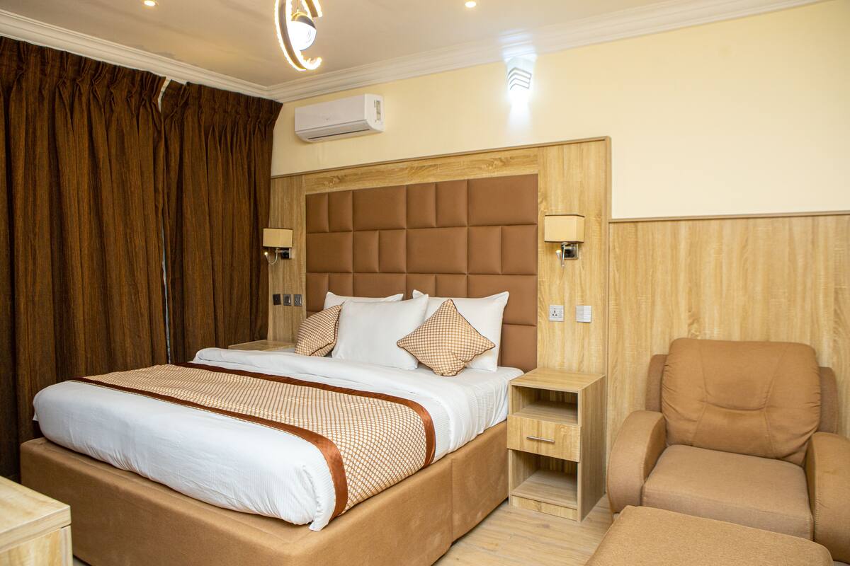 Habitación de hotel con una cama grande, cabecera de madera, dos mesitas de noche, una silla y un aire acondicionado.