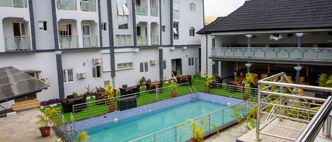 A swimming pool surrounded by potted plants, a covered seating area, and a multi-story building with balconies.