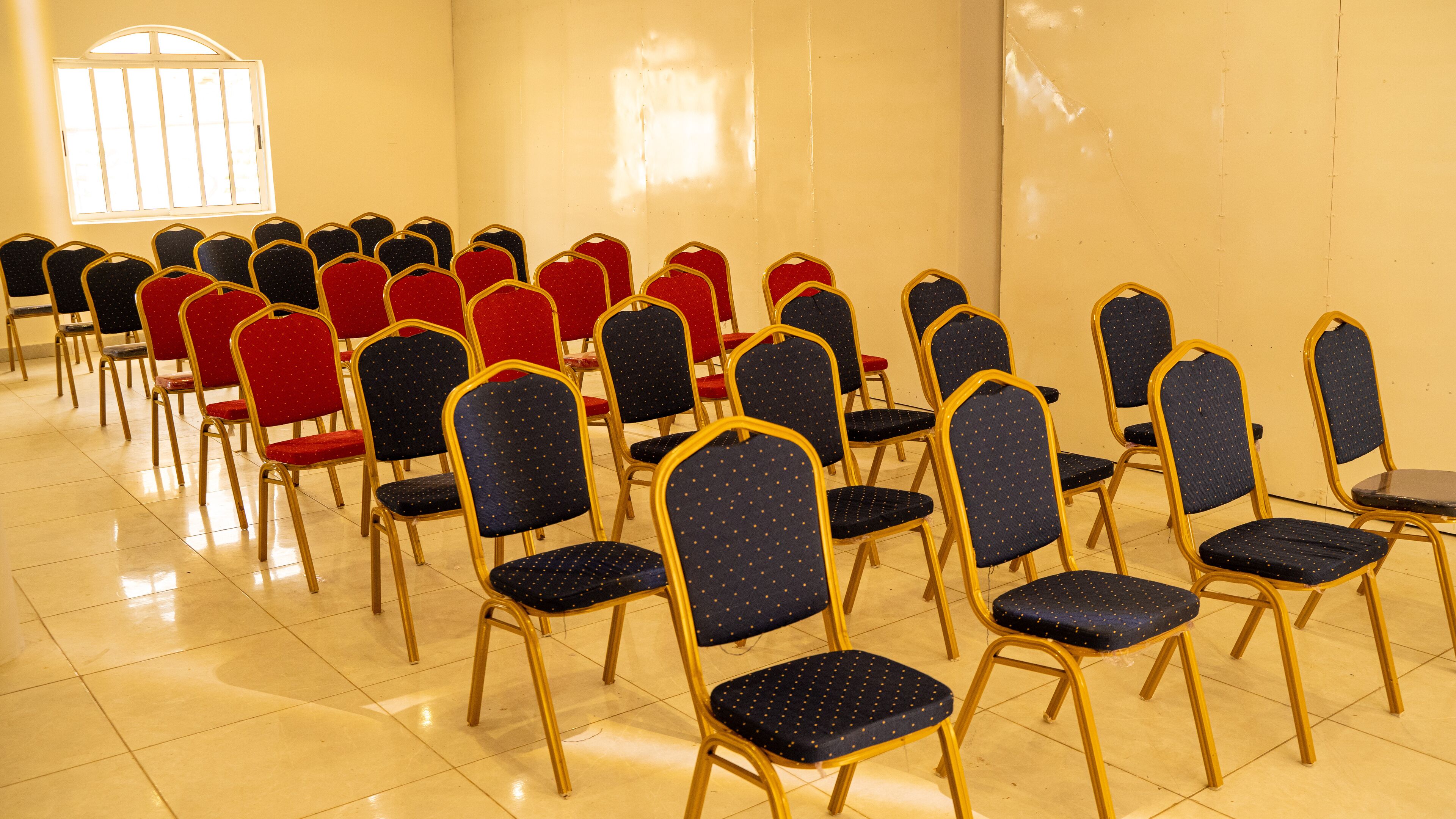 Rows of foldable chairs with red and blue upholstery in a room with a tiled floor and a window with bars.