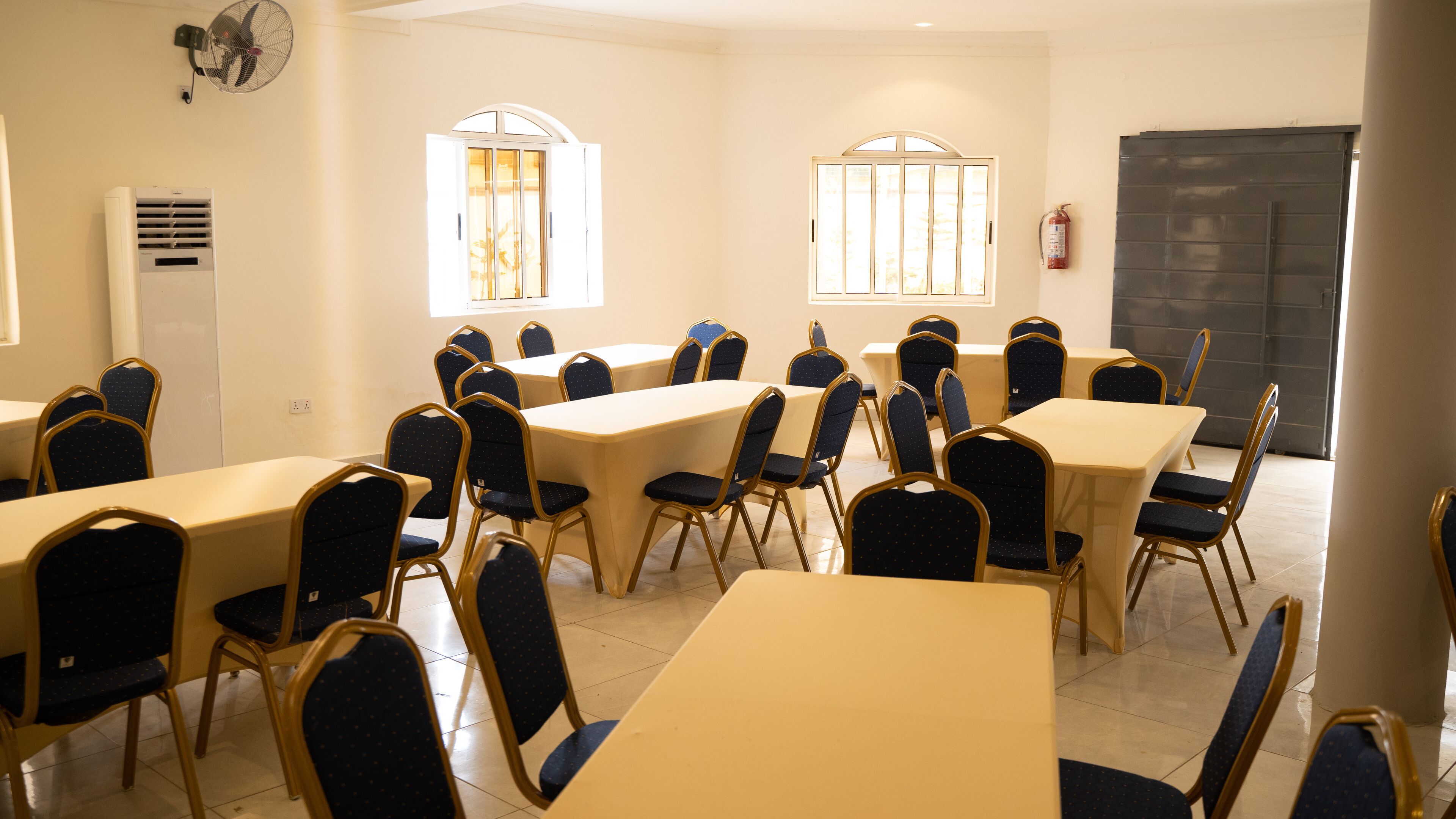 Una sala conferenze con diversi tavoli e sedie, ampie finestre e un estintore appeso al muro.