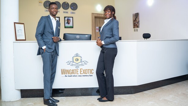 A reception desk with a logo for Wingate Exotic hotel, featuring a clock, framed photos, and a computer. - Wingate Exotic (Ilorin)