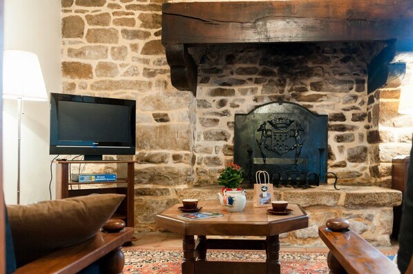 A stone fireplace with a decorative metal plaque, a television on a wooden stand, and a table with a teapot and cups. - Beautiful HOUSE in a quiet area ***—Ocean: 20 min—Quimperlé-Le Faouet: 10 km—with a SPECIAL OFFER (QUERRIEN)