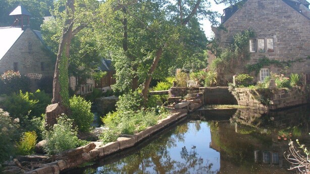 Ein Steingebäude mit einer kleinen Brücke über einem ruhigen Wasserlauf, umgeben von üppiger Vegetation und Bäumen.