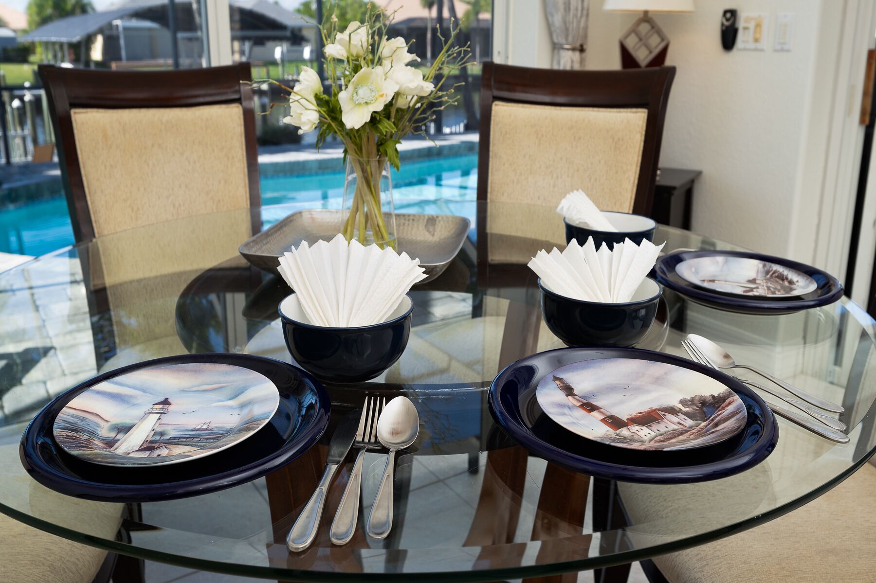 Une table ronde en verre, placée devant une piscine, entourée de chaises et dressée avec des assiettes, des bols et des couverts.